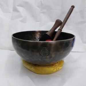 May include: A dark gray metal singing bowl with a decorative design on the bottom. The bowl is sitting on a yellow cushion and has two wooden mallets with red-tipped ends.