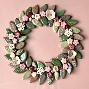 Peut inclure: Une couronne circulaire composée de nombreuses feuilles et fleurs en céramique. Les feuilles sont émaillées dans diverses nuances de vert et de brun, tandis que les fleurs sont réalisées dans des tons de blanc, rose pâle et rouge foncé.