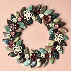 May include: A circular wreath composed of individual ceramic pieces shaped like leaves and flowers. The leaves are in shades of green, brown, and teal. The flowers are in cream, dark red, and pink, featuring glossy finishes and varied textures.