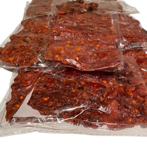 May include: Close-up of several clear plastic packages filled with red-brown jerky. The jerky has a slightly glossy appearance and is speckled with red pepper flakes. The packages are stacked on top of each other, with some overlapping.