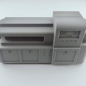 Puede incluir: Modelo gris impreso en 3D de una computadora con las letras "WOPR" en el lateral. El modelo tiene una base rectangular con dos puertas tipo armario y una sección superior con un diseño similar a una rejilla.