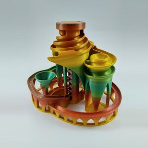 May include: A colorful 3D printed marble run with a tower and multiple levels. The run is made of orange, yellow, green, and red plastic.