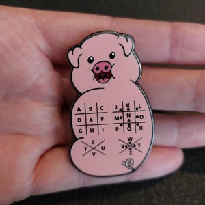 May include: Pink enamel pin shaped like a pig with a braille alphabet on its back. The braille alphabet is arranged in a grid with letters A through Z.