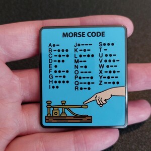 May include: A blue and black enamel pin with a Morse code chart and a cartoon illustration of a hand operating a telegraph key. The text on the pin reads "MORSE CODE".