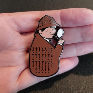 May include: A brown enamel pin depicting a cartoon detective wearing a brown hat and coat, holding a magnifying glass. The detective is looking through the magnifying glass at a chart of letters and symbols. The chart includes the letters A through Z and symbols for each letter.