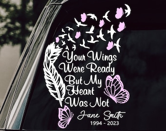 May include: A white and pink memorial decal on a car window. The decal features a feather, butterflies, and birds, with the text "Your Wings Were Ready But My Heart Was Not" and a name and dates.