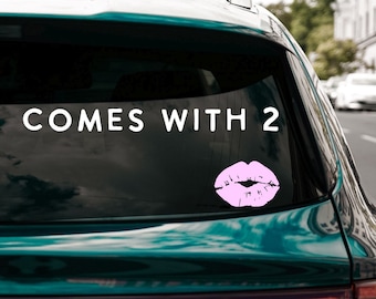 Puede incluir: Una calcomanía para ventana de coche con el texto "COMES WITH 2" en blanco, acompañada de un gráfico de labios rosas. La calcomanía está adherida a la ventana trasera de un coche verde oscuro, con una calle de la ciudad al fondo.