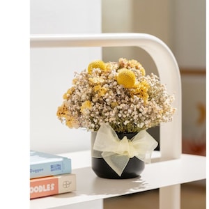 May include: A black vase with a yellow bow holds a bouquet of dried flowers. The flowers are a mix of small white flowers and larger yellow pom pom flowers.