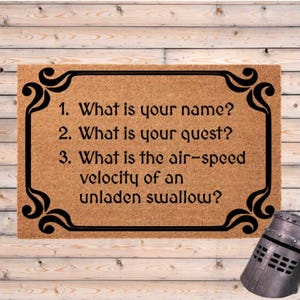 May include: A rectangular doormat with a light brown background and black decorative border. The text reads: "1. What is your name? 2. What is your quest? 3. What is the air-speed velocity of an unladen swallow?" A knight's helmet is in the corner.