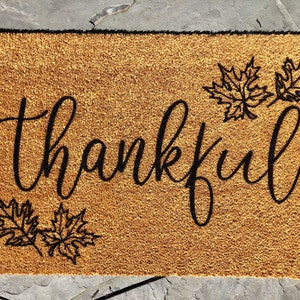 K&ouml;nnte beinhalten: Eine rechteckige Fu&szlig;matte aus nat&uuml;rlicher Kokosfaser. Auf der Matte steht das Wort "thankful" in schwarzer Schrift, zusammen mit drei schwarzen Ahornblatt-Designs. Die Matte ist f&uuml;r den Au&szlig;enbereich konzipiert und liegt auf einer Steinoberfl&auml;che.