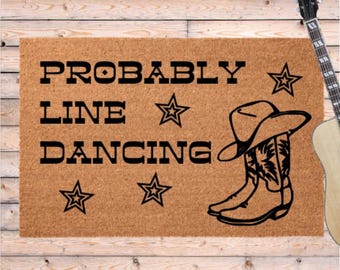 May include: A rectangular doormat with the words "PROBABLY LINE DANCING" in black, along with star and cowboy boot graphics. The mat is a natural tan color, and the design is simple and bold. A guitar is in the background.