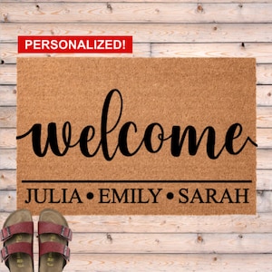 May include: A rectangular doormat made of natural coir fiber, featuring the word "welcome" in elegant black script. Below, the names "JULIA • EMILY • SARAH" are printed. The word "PERSONALIZED!" is in a red box. Red sandals are in the foreground.