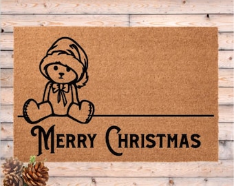Könnte beinhalten: Rechteckige Fußmatte mit brauner Oberfläche. Auf der linken Seite befindet sich eine schwarze Umrisszeichnung eines Teddybären mit Weihnachtsmütze. Die Worte "Merry Christmas" sind schwarz geschrieben.