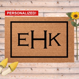 May include: A rectangular doormat with a black border and the letters "EHK" in the center. The word "PERSONALIZED!" is in a red rectangle in the upper left corner. The mat is brown, and the background is a wooden surface.