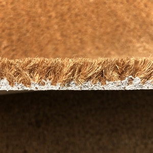 May include: Close-up of a brown coir doormat with a white backing. The coir fibers are visible, creating a textured surface. The doormat is likely used for wiping shoes at an entrance.