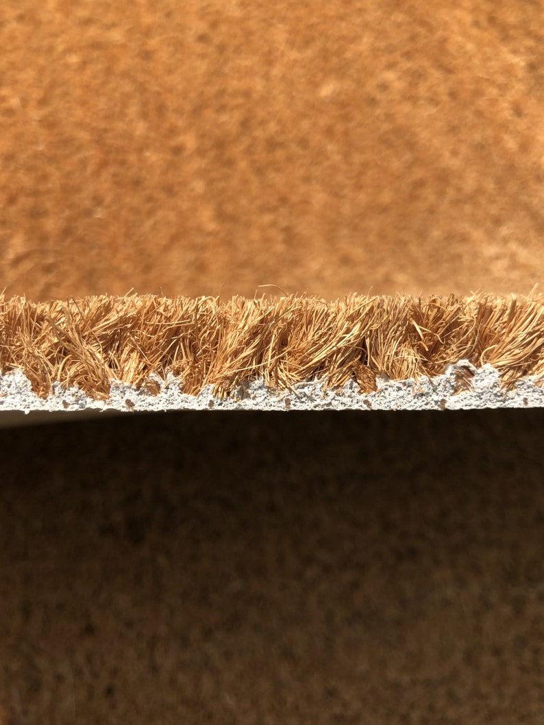 May include: Close-up of a brown coir doormat with a white backing. The coir fibers are visible, creating a textured surface. The doormat is likely used for wiping shoes at an entrance.