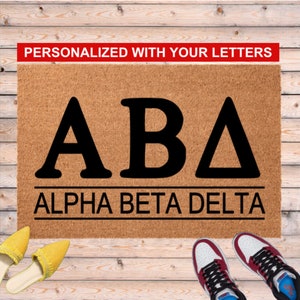 May include: A rectangular doormat made of natural coir fiber. The mat features the letters "ABA" in bold black font, with "ALPHA BETA DELTA" below. The text "PERSONALIZED WITH YOUR LETTERS" is in a red banner at the top.