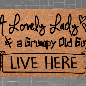 May include: A rectangular doormat made of natural coir fiber. The mat features black text that reads "A Lovely Lady & a Grumpy Old Guy LIVE HERE", with heart symbols. The design is centered on the mat.