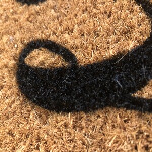 May include: Close-up of a brown coir doormat with a black, stylized letter. The mat has a textured surface and is designed for outdoor use. The black letter is a bold, decorative element.