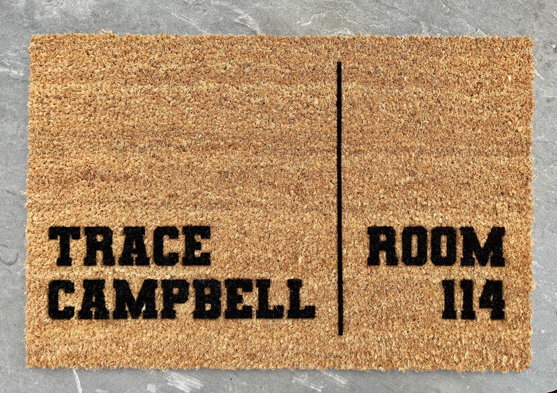 May include: A rectangular doormat made of natural coir fibers. The mat features the name "TRACE CAMPBELL" on the left and "ROOM 114" on the right, separated by a vertical black line. The text is in a bold, black font.