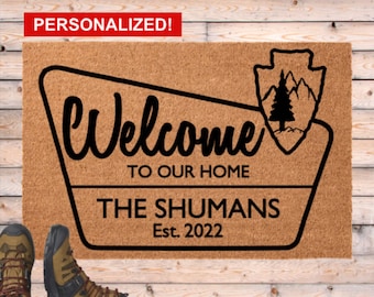 Könnte beinhalten: Eine rechteckige Kokos-Fußmatte mit den Worten "Welcome to our Home" in schwarzer Schrift. Darunter steht "The Shumans Est. 2022". Eine schwarze Grafik eines Berges und eines Baumes befindet sich rechts. Das Wort "PERSONALISIERT!" steht in einem roten Feld.