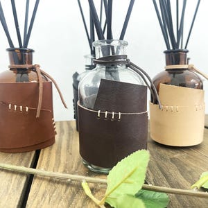 May include: Several glass diffuser bottles with black reeds. Each bottle has a leather sleeve, stitched with white thread. The leather sleeves are in shades of brown and tan, creating a rustic aesthetic.