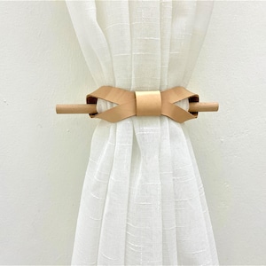 May include: A light brown leather curtain tie with a wooden dowel. The tie is tied in a bow shape and is holding back a white sheer curtain.