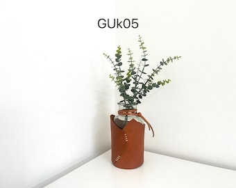 May include: A clear glass vase with a brown leather sleeve and green leafy stems. The leather sleeve is stitched with white thread and has a strap. The text "GUK05" is at the top.