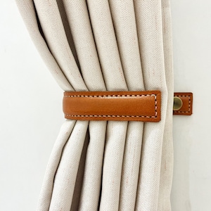 May include: A brown leather curtain tieback with a stitched edge and a metal buckle. The tieback is holding back a set of white curtains.