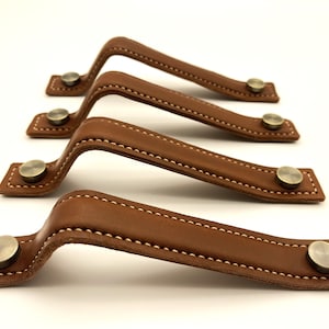 May include: Four brown leather cabinet pulls with brass accents. Each pull has a stitched edge and a rounded shape.