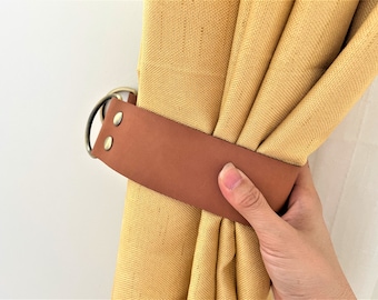 May include: A brown leather curtain tieback with two gold metal rings. The tieback is holding back a yellow curtain.