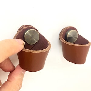 May include: Two brown leather cabinet pulls with a brass knob in the centre. The pulls are attached to a white wall.