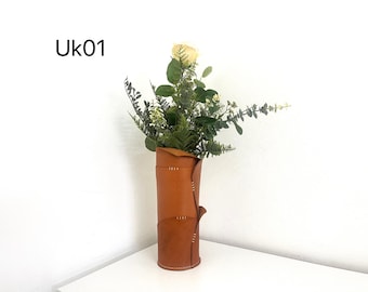 May include: A tall, cylindrical brown leather vase with a rolled top, stitched details, and a decorative flap. The vase holds a bouquet of greenery and a single cream-colored rose. The text "Uk01" is in the upper left corner.