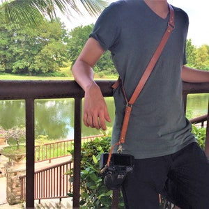 May include: A person wearing a teal t-shirt and black trousers with a brown leather camera strap. A camera is attached to the strap.