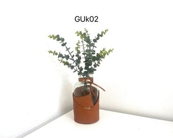 May include: A clear glass vase with a brown leather sleeve and a bundle of green eucalyptus stems. The vase is tied with a leather cord. The text "GUK02" is in the top left corner.
