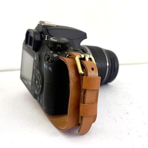 May include: A black DSLR camera with a brown leather strap and a silver buckle. The camera has a lens attached and a screen on the back.