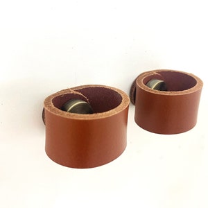 May include: Two brown leather rings with a brass metal accent. The rings are designed to be used as a belt loop or a key chain.