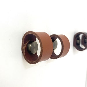 May include: Two brown leather cabinet pulls with a brass screw in the center of each pull. The pulls are attached to a white wall.