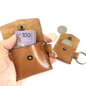 May include: A brown leather keychain with a small pocket for cash or coins. The keychain has a metal ring and a snap closure. The pocket is open and a bill with the text "RM 100" is visible.