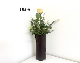May include: A tall, dark brown leather vase with a cream-colored rose and assorted greenery. The vase has a stitched design and the text "UK05" is in the upper left corner. The arrangement includes eucalyptus and other green foliage.