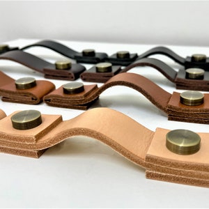 以下が含まれることがあります： 真鍮製の金具が付いた革製のキャビネットプルをいくつかクローズアップで写した写真です。プルは、薄い茶色、濃い茶色、黒など、さまざまな茶色の色合いです。