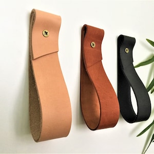 May include: Three leather wall hooks in different colors: light brown, brown, and black. Each hook is a single piece of leather with a screw in the top.