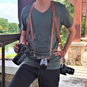 Puede incluir: Una persona que lleva una camiseta verde y correas de cámara de cuero marrón con dos cámaras conectadas. Las cámaras son una cámara réflex digital negra con un objetivo y una cámara sin espejo plateada con un objetivo.