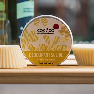 Peut inclure: Un pot rond de déodorant solide avec un design à rayures jaunes et blanches. L'étiquette indique "coclico Cosmetiques Naturels DEODORANT SOLIDE Noix de coco Fait à Paris avec ♥". Deux bâtonnets de déodorant blancs sont également visibles.