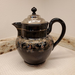 May include: A black ceramic teapot with a silver lid and a floral design. The teapot has a gold trim and a black handle.