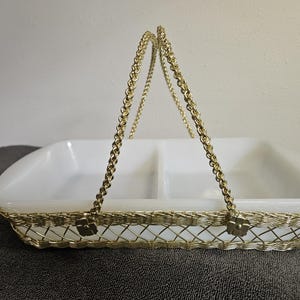 May include: A white divided serving dish with a gold-coloured wire basket and chain handle. The basket has a woven design, and the handle is made of a gold chain. The dish is rectangular with rounded corners.