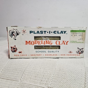 Pode incluir: Caixa vintage de plasticina Plast-I-Clay. A caixa branca apresenta ilustrações de um elefante, um gato e flores. O texto "MODELING CLAY" é vermelho. A caixa está rotulada como "SCHOOL QUALITY" e "NON-TOXIC".