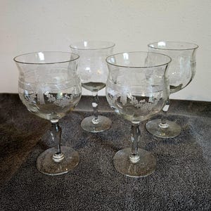May include: A set of four clear glass goblets featuring a delicate etched pattern around the bowl. Each glass has a smooth, rounded rim, a curved bowl shape, a slender stem, and a circular base. The etched design includes ribbons and floral motifs.