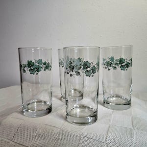 May include: Four clear highball glasses with a green and blue leaf design around the top. The glasses are cylindrical and sit on a white tablecloth. The glasses are empty.