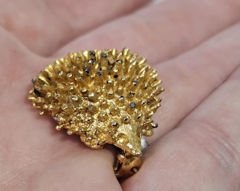 May include: A gold-colored hedgehog ring with a textured surface and small dark accents. The ring features a detailed design of the hedgehog's quills and face, showcasing a unique and whimsical style. The ring is a statement piece.